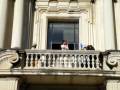 Dos personas en un balcón de un edificio histórico, uno con una gorra y el otro con una cámara, posiblemente fotografiando o grabando algo.
