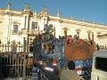Desfile de carros alegóricos en una plaza histórica, con una estructura arquitectónica imponente al fondo.