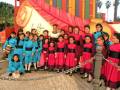 Un grupo de niñas vestidas con trajes azules y rojos posan frente a un escenario colorido, con una figura de la Virgen en el fondo.