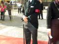Un hombre vestido formalmente con un traje verde y sombrero negro, posa en un entorno urbano con otros individuos vestidos de manera similar. La imagen sugiere una celebración o evento formal, posiblemente un desfile o ceremonia.