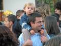 Una familia en un ambiente festivo, con una niña pequeña sentada en los hombros de su padre. El padre sonríe, mientras la niña mira hacia el lado. La imagen muestra a varias personas de diferentes edades y estilos, en un entorno que parece ser una celebración o evento público.