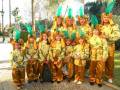 Un grupo de niños disfrazados con trajes verdes y amarillos, vestidos como nativos americanos. Ellos llevan sombreros con plumas y pantalones largos de color marrón. La escena está en un parque con árboles y una calle de fondo.