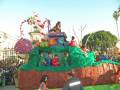 Un carnaval con un carro de fantasía de Halloween, decorado con calabazas y una figura de calabaza en un trono. La imagen muestra a niños vestidos como fantasmas y una figura de calabaza en un trono. La imagen también muestra a personas observando el carro desde los lados.