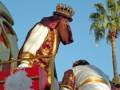 Un personaje vestido con trajes de estilo medieval, incluyendo una corona y un manto rojo y dorado, se encuentra en el centro de la imagen. A su lado, hay una estructura que parece ser parte de un carro o carreta tradicional. En el fondo, se pueden ver árboles y una estructura que podría ser parte de un palacio o edificio histórico. La imagen sugiere un evento festivo o ceremonial, posiblemente relacionado con la cultura medieval.