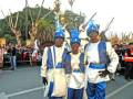 Tres personas vestidas con trajes tradicionales participan en un desfile. La imagen muestra a tres individuos vestidos con trajes azules y blancos, con sombreros decorativos. La escena se desarrolla en una calle con árboles y espectadores observando el desfile.