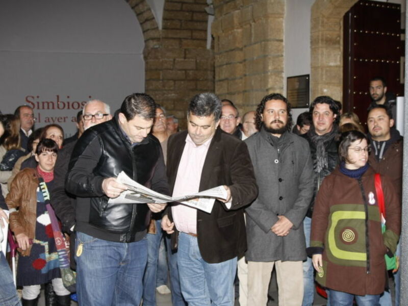 Persona leyendo un periódico mientras una multitud observa en un espacio interior con arcos y columnas.