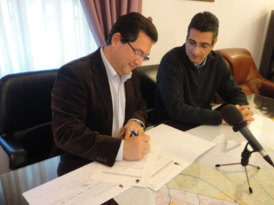Dos hombres sentados en una oficina, uno con un traje y el otro con una sudadera, firmando documentos junto a una mesa con un mapa en la pared.