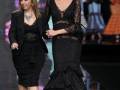 Dos modelos caminan por la pasarela en un desfile de moda, uno con un vestido negro de corte sirena y el otro con un vestido negro ajustado. La escena está iluminada por una pantalla de fondo que muestra un diseño rojo y blanco.