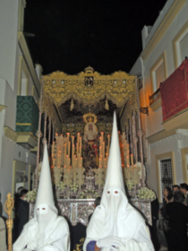 Una procesión religiosa nocturna con penitentes vestidos de blanco y sombreros largos, frente a una iglesia adornada con velas encendidas.
