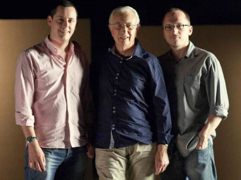 Tres hombres posando juntos en una sala de conferencias.