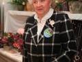 Una mujer con cabello blanco y una chaqueta a cuadros negra y blanca. Detrás de ella hay flores y una mesa con mantel blanco.