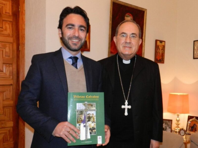 Un hombre en traje sostiene un libro mientras está junto a otro hombre con una cruz. El ambiente parece ser formal, posiblemente en un entorno religioso o institucional.