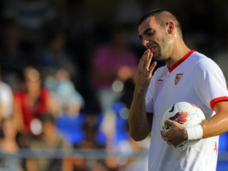 Un jugador de fútbol sostiene una pelota mientras se prepara para un partido. La escena está enfocada en el jugador, con el resto del campo y a los espectadores en el fondo.