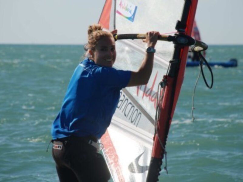 Un windsurfer disfruta de un día soleado en el mar, sujetando firmemente el mástil de su vela roja y blanca.