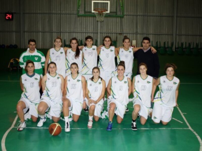 Equipo femenino de baloncesto posando en el campo de juego, con uniformes blancos y verdes, y una canasta en el fondo.