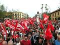 Una multitud de personas con banderas rojas y blancas, posiblemente en una manifestación política o sindical. Fondo urbano con edificios y árboles en la distancia.