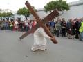 Una persona vestida como Jesús lleva una cruz en la cabeza, mientras otros participantes observan desde el lado. La escena parece ser parte de una representación o acto religioso en un entorno urbano.