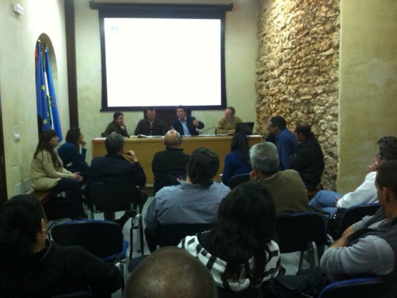 Una sala de conferencias con una pantalla proyectora en el centro. Un hombre está hablando a un grupo de personas sentadas en sillas. La sala tiene una pared de piedra en el fondo y una bandera colgada en la pared izquierda.