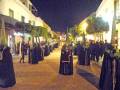 Una procesión nocturna en una calle adoquinada, con personas vestidas de negro y capas largas. La iluminación proviene de farolas y luces en los edificios, creando una atmósfera festiva.