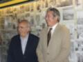 Dos hombres sonrientes frente a una pared de fotos antiguas. El ambiente parece ser un lugar público o museo con numerosas imágenes enmarcadas.
