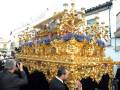 Una procesión religiosa con un carro de oro dorado y figuras talladas, adornado con flores azules. La imagen muestra a personas observando y fotografiando el evento.