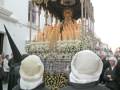 Una procesión religiosa con una imagen de la Virgen en un carro decorado con flores y velas. Dos personas con gorros blancos y chaquetas negras con letras en español. Fondo de edificios blancos.