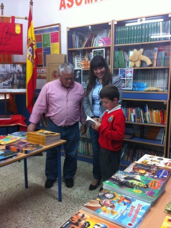 En una biblioteca, un hombre y una mujer observan con atención a un niño que sostiene un libro. La escena está rodeada de estanterías llena de libros y una mesa con más títulos.