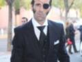 Un hombre viste un traje negro con detalles en la manga y una corbata negra. Lleva una camisa blanca con un pañuelo en el bolsillo de la solapa. Su rostro muestra una cicatriz y un ojo negro con una venda, sugiriendo un personaje de ficción.