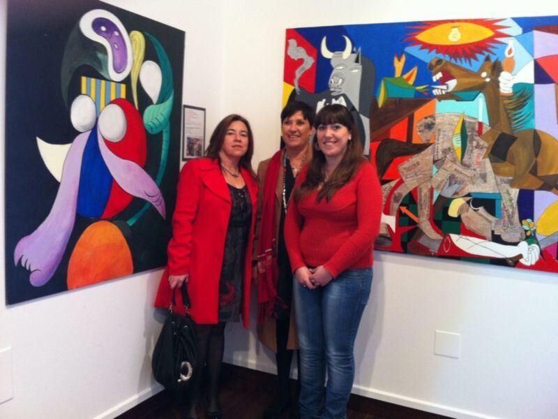 Tres personas posan frente a dos obras de arte vibrantes en una exposición. La imagen muestra un ambiente cultural y creativo.