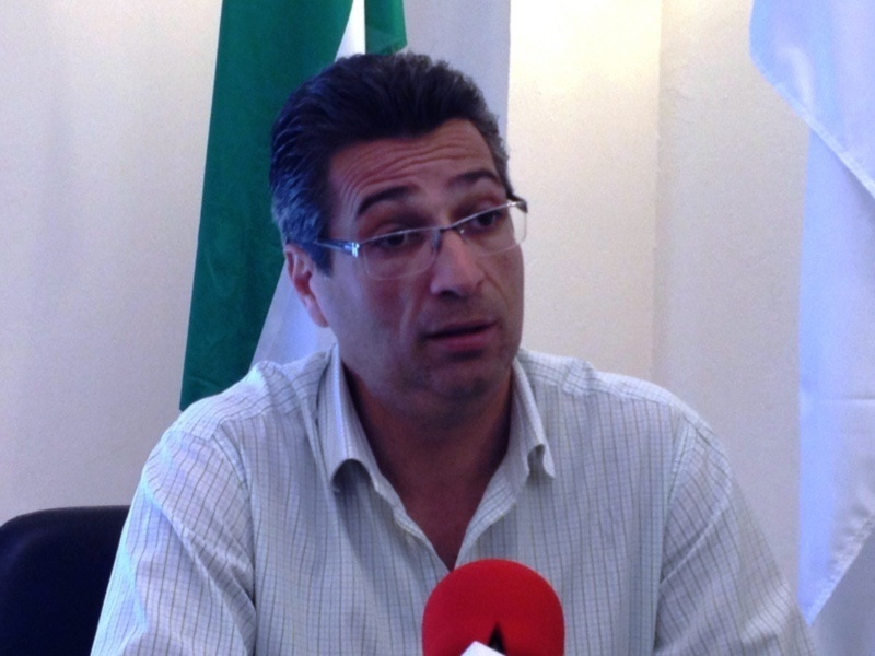 Un hombre con gafas hablando en una conferencia de prensa, con un micrófono rojo frente a él y banderas en el fondo.