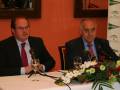 Dos hombres en una conferencia de prensa con microfonos y copas de vino en una mesa decorada con flores.