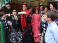 Una mujer en traje de flamenca roja y negra se prepara para cortar una tarta. A su alrededor, personas de diferentes edades observan la escena con interés. La imagen se desarrolla en un ambiente festivo, posiblemente durante una celebración tradicional española.