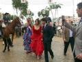 Una mujer en un vestido rojo flamenco camina junto a un hombre en traje, mientras otros espectadores observan. A la izquierda, caballos y personas montadas en ellos se destacan contra un fondo de arcos decorativos.