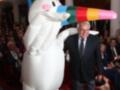 Un hombre en traje negro sonríe mientras sostiene un unicornio inflable blanco con arcoíris en la cabeza. La imagen se toma en un evento formal, con asistentes sentados en el fondo y luces de teatro.
