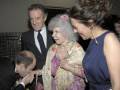 Una anciana con cabello blanco y una camisa floral se abraza con un hombre en traje negro. A su lado, una mujer joven en vestido azul sostiene a la anciana. La escena parece ser una celebración o evento formal, con un fondo de luces y decoraciones.