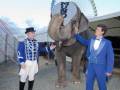 Dos personas en trajes de circo posan con un elefante. En el fondo, se ve una gran rueda de ferris y un puente.