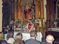 Una imagen de una iglesia con un altar dorado y una estatua central. Un sacerdote en rojo bendice a los fieles sentados en bancos de madera. La atmósfera es reverente y religiosa, reflejando una celebración o misa.