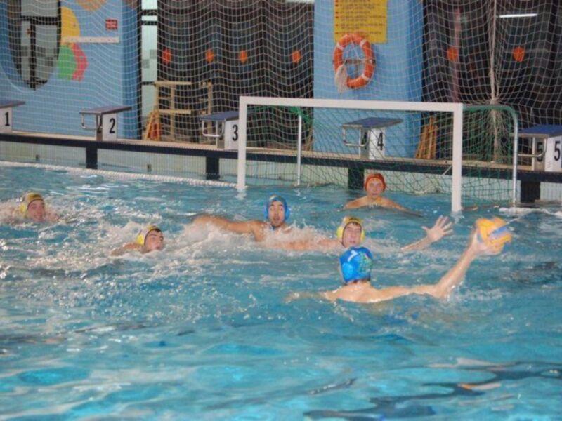 Jugadores de waterpolo en acción en una piscina interior, con un portero en el centro y otros jugadores alrededor, preparándose para el lanzamiento del balón.