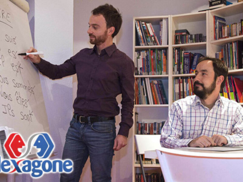 Un hombre está escribiendo en una pizarra mientras otro lo observa. Ambos están en un entorno de oficina con estanterías llenas de libros y material. La imagen muestra la marca Hexagone en el fondo.