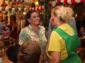 Dos mujeres charlan en un ambiente festivo con decoración navideña. Una mujer con peluca amarilla y otro niño están cerca de ellas. La decoración incluye globos rojos y blancos, una araña grande en el centro y luces colgantes. La imagen transmite un ambiente festivo y familiar.