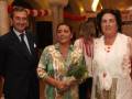 Una familia en un evento formal, con una mujer en el centro sosteniendo flores, rodeada de dos personas, posiblemente su marido y su madre, todos sonrientes y vestidos de manera elegante.