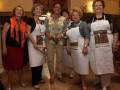 Seis mujeres sonrientes posan con delantales en un ambiente festivo. La imagen muestra una celebración o evento social, posiblemente relacionado con la gastronomía.
