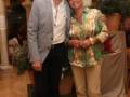 Una pareja posa juntos en un ambiente festivo decorado con globos y flores. La persona de la izquierda lleva un traje gris claro, mientras que la de la derecha usa una camisa con estampado floral y pantalones beige. Ambos parecen estar en un evento celebrativo.