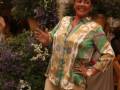 Una mujer con un vestido de flores brillantes y una corona sonríe ampliamente, rodeada por flores en un ambiente festivo.