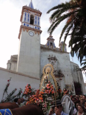 Fiesta religiosa con una imagen de la Virgen en un carro, frente a una iglesia con torres y un reloj.