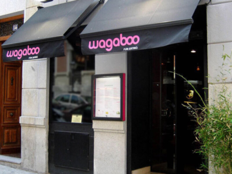 Entrada de un restaurante con una puerta de madera y un letrero de acero inoxidable con el nombre 'wagaboo' en letras rosas y negras.