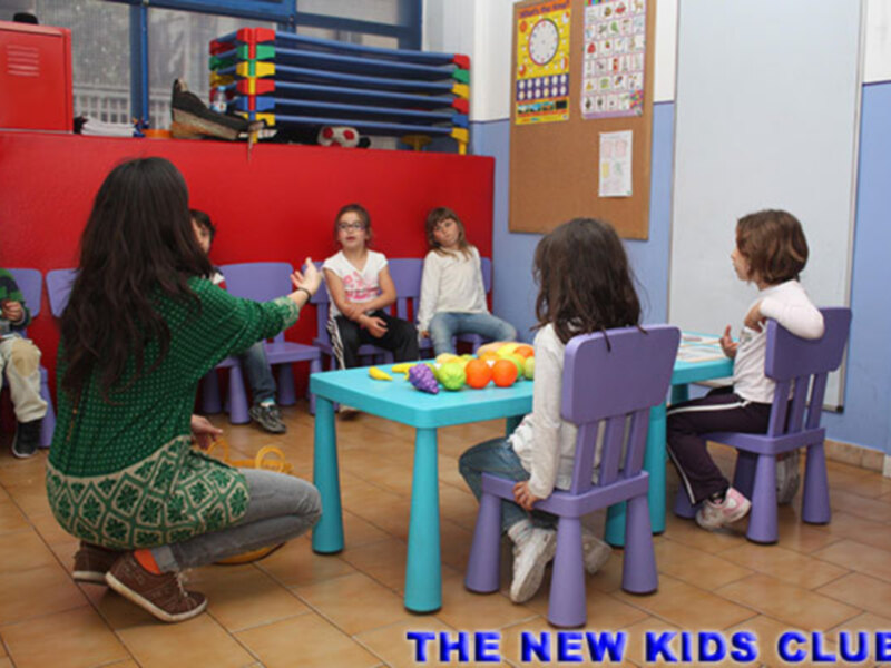 Niños en una escuela infantil, sentados alrededor de una mesa azul con frutas y juguetes. Una maestra se inclina para interactuar con ellos. En la pared hay un calendario y una lista de actividades. La imagen está firmada "THE NEW KIDS CLUB".
