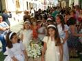 Fotografía de una procesión religiosa en un pueblo español con niñas vestidas de blanco y flores, gente en la calle y edificios típicos.