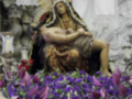 Una figura de la Virgen María sentada en un trono, rodeada por flores y adornos. La escena parece ser parte de una celebración religiosa o procesión, con un fondo que sugiere un ambiente sacro.