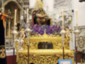 Una estatua de oro en un altar dorado, rodeada por velas y decoraciones. En el fondo, se puede ver una ventana con vidrieras. Al lado derecho hay un cartel informativo azul sobre el altar.